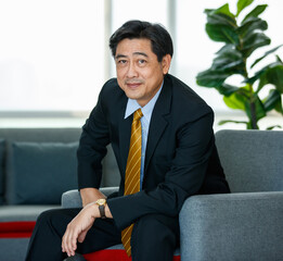 50s Asian senior executive businessman entrepreneur wearing formal suit with luxury necktie, smart sitting on sofa in indoor office, smiling with confidence, success and gladness with copy space.