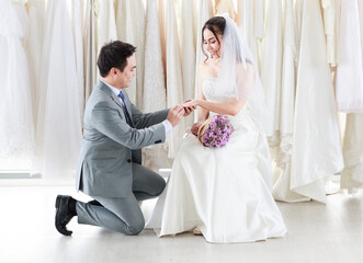 The groom in a gray suit kneels and wears a wedding ring to the bride in white lace in the room at studio. Asian couples having a blessed best day to share life. Concept happy lovely love.