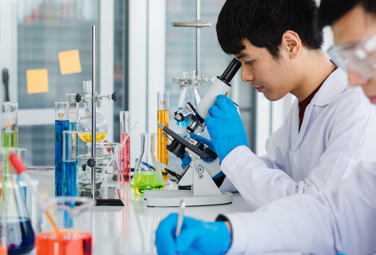 Asian young man sitting and looking through microscope and adult man wearing eyeglass do writing a note book both are chemist scientist and in uniform.Test tube with liquid put on table in laboratory