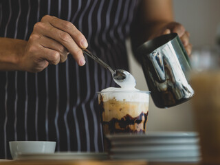 Unrecognizable  barista using spoon to gentle topping glass of sweet coffee at bar counter with white milk froth from metal jar to enhance taste of softness and mellowness.