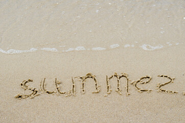 The word SUMMER on the yellow sand. Handwritten word on a wet sandy beach. View from above at an angle.