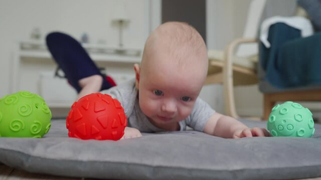 Cute baby lying on his tummy on play mat, 3 month old newborn infant boy playing with colorful sensory balls on the floor. High quality 4k footage