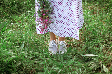 the legs of a beautiful girl in a long white dress and gray shoes are standing in the tall green grass. A girl holds a bouquet of purple wildflowers. Selective focus. Copy Space