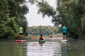 Two guys on SUP and a girl in a kayak row on the Danube near green trees