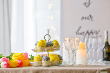 Dessert stand with tasty cupcakes, burning candles, glasses and flowers on table