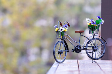 A decorative metal forged tricycle with a basket holding plastic flowers standing on a wooden deck with blurry background,  Sunriver, Oregon