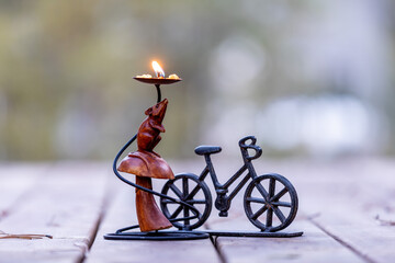 An isolated wooden mouse doll under a drip pan of a decorative burning candle stand against blurry background. Figure of a mouse standing on a back paws under an umbrella next to a toy metal bike