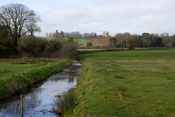 Fototapeta na wymiar drainage channel in typical English farmland with Folly in the distance and a blue sky and white clouds