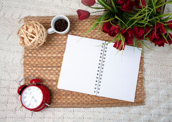 An open notebook, an alarm clock, and a cup of coffee on flowers on the bed. Place for your lettering, drawing, logo	