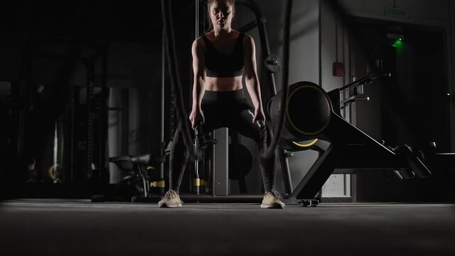 Caucasian athletic female fighter performs exercises with battle ropes, endurance training in the gym.