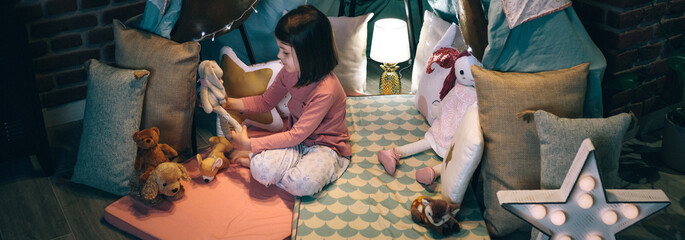 Adorable girl playing alone with teddies in a tent made of bed sheets and chairs