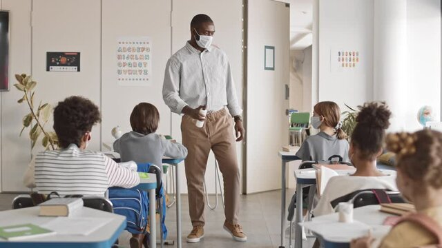 Tracking slowmo of responsible African-American school teacher in smart casualwear and face mask walking along rows in classroom disinfecting hands of his students sitting at desks with sanitizer