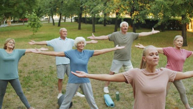 Zoom in shot of group of elderly men and women in sportswear doing stretching exercise in park while having outdoor workout with female coach