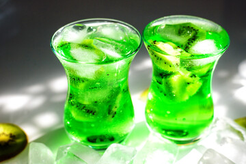 Green lemonade with ice and kiwi slices.Two glass glasses stand on the table and the sun's rays fall on them.
