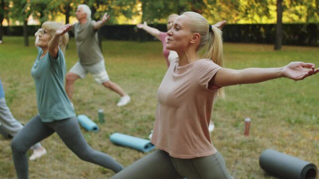 Tilt up shot of group of elderly people in sportswear standing in lunge and raising arms up while practicing yoga outdoors in park with female instructor
