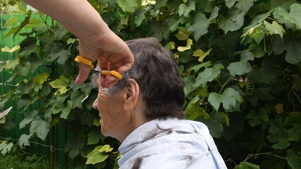 Woman hands trim hair on temple of senior person with half gray haired short haircut. Female hands...