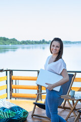 Young woman with closed net-book laptop standing outdoor, next to the river holding laptop computer in hand while resting outdoors ready to work. Remote job