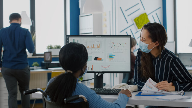 Freelancers wearing protective face masks working on computer in business office during global pandemic, disabled african woman sitting in wheelchair . Team discussing in new normal financial office