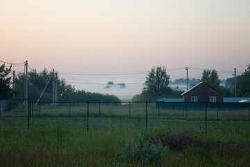 A village with a wooden house and a fence in the early foggy summer morning at dawn