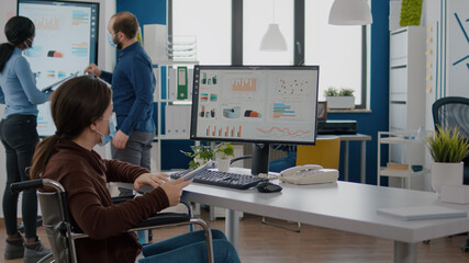 Handicapped invalid businesswoman with protection mask against covid19 pandemic analysing financial statistics sitting in wheelchair, checking graphs from pc in new normal start up business office.