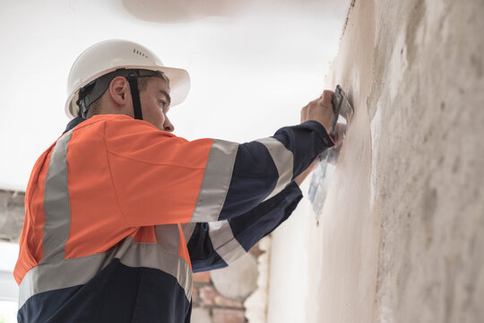 A man in a helmet and a work uniform aligns the walls under the ceiling with a spatula and putty.