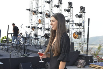Fototapeta na wymiar Happy young woman smiling and drinking wine. Winery event. Young girl portrait. Stage and music background. Black outfit. Event in the winery