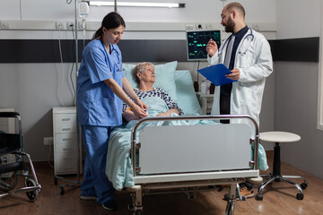 Medical nurse attaching oxymeter on senior woman patient, laying on hospital bed discussing with doctor about diagnosis and treatment while she breaths with help from oxygem mask.