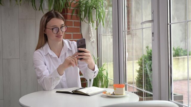 Caucasian blond woman in glasses sits in a cafe and uses her smartphone to make a video call.