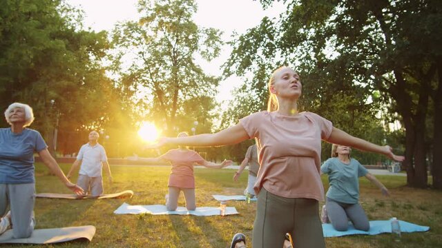 Arc shot of young female teacher speaking and holding Namaste while leading outdoor yoga class with elderly people in park at sunset
