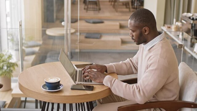 Medium side-view shot of modern African-American man in smart casualwear working on laptop at cafe table