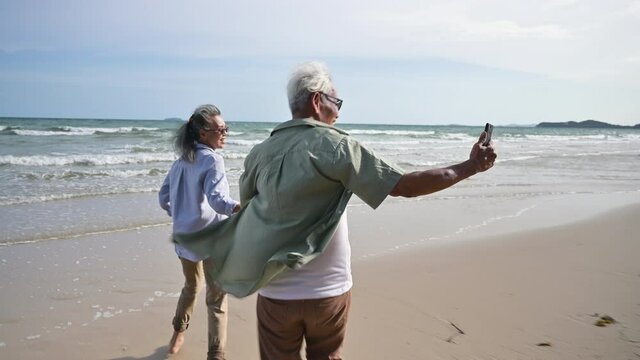Asian senior woman wear sunglasses smile and hand holding husband running from beach to sea in vacation. Happy family couple elderly older retire shooting video and selfie by smartphone on seaside. 