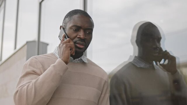 Medium shot of modern African-American businessman in smart casualwear making phone call standing by glass office building
