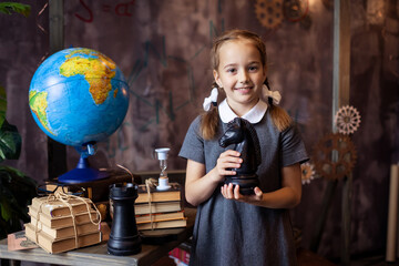 A smart schoolgirl holds chess pieces in her hands. The girl with pigtails is a chess player. Primary School. Development of mind and logic. Little genius. The globe. Workplace of a school child