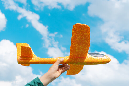 Child hand holding cheerful orange toy plane on blue sky background. Happy childhood, freedom and aspirations concept.