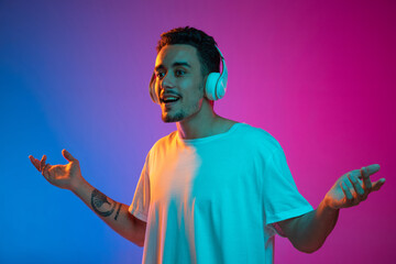 Portrait of Latina young man in white headphones listening to music isolated on gradient purple pink background in neon light.