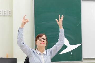 cute young woman teacher at school at her workplace sitting at the table scatters documents on the background of the blackboard