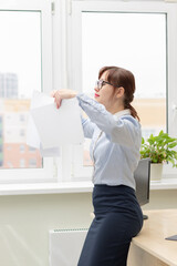 pretty young woman worker in the office at her workplace standing by the table in front of the window scatters documents