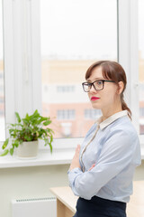 young pretty business woman in glasses and a blue blouse stands at the desktop in the office against the background of a light window