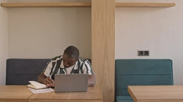 Medium front-view shot of young African-American male freelancer in casualwear witting at cafe table with laptop taking notes in copybook