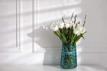 Beautiful bouquet of willow branches and tulips in vase near white wall, space for text