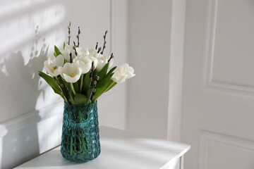 Beautiful bouquet of willow branches and tulips in vase near white wall indoors, space for text