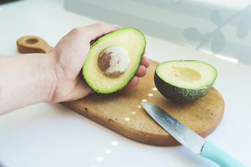 Male caucasian hand holding a half cut haas avocado on a wooden kitchen board with a knife. Photo for step by step avocado salad recipes. The concept of cooking, proper healthy nutrition, vitamins.