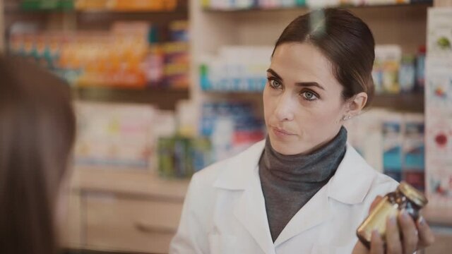 A serious professional pharmacist woman is consulting and talking how to use a drug capsule in a pharmacy standing at the cashbox