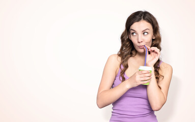 Young happy girl drinking through a straw from a paper cup and looking away, on a light yellow background 