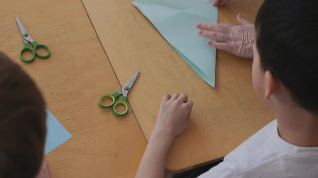 The Hands Of An Elderly Teacher Makes Shapes Out Of Paper With Children Sitting At The Table. Top View. Preschool Education.