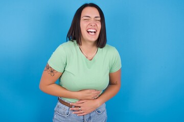 young beautiful brunette girl with short hair standing against blue background smiling and laughing hard out loud because funny crazy joke with hands on body.