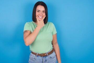 Fototapeta na wymiar young beautiful brunette girl with short hair standing against blue background looking confident at the camera smiling with crossed arms and hand raised on chin. Thinking positive.