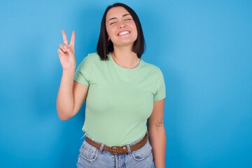 young beautiful brunette girl with short hair standing against blue background smiling with happy face winking at the camera doing victory sign. Number two.