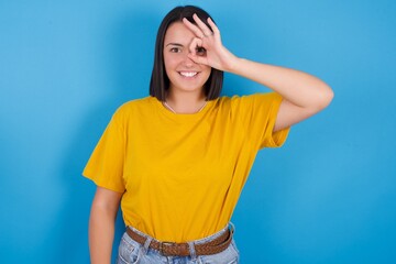 young beautiful brunette girl with short hair standing against blue background doing ok gesture with hand smiling, eye looking through fingers with happy face.