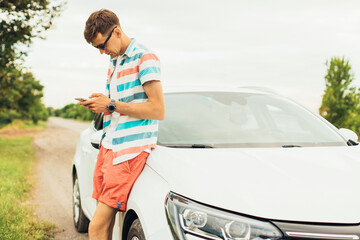young man in summer clothes and sunglasses standing near his luxury white car and using smartphone, holding in hand